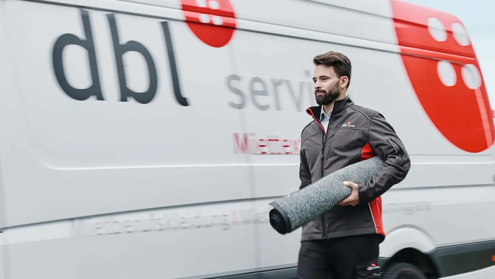 Das Bild zeigt einen DBL-Servicefahrer, der eine saubere Schmutzfangmatte zu einem Unternehmen liefert. Der Mietservice sorgt für regelmäßigen Austausch und gewährleistet so eine effektive Schmutz- und Keimabwehr in den Eingangsbereichen, besonders in der nassen Winterzeit. 
